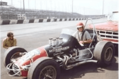 A person in a vintage race car on a racetrack, wearing a helmet, with another person crouched nearby. A truck is parked in the background.