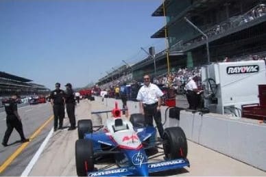 A person stands next to a race car in the pit lane of a racetrack, surrounded by teams and spectators.