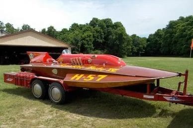 Red speedboat labeled "H-57" on a trailer, parked on grass near a building and trees.