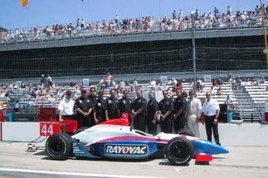 A racing team poses beside a Rayovac branded race car on the track with a large crowd in the grandstand behind them.