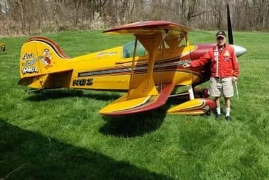 A man stands next to a small yellow and red vintage biplane on a grassy field.