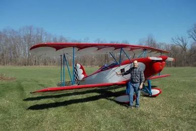 A man stands next to a red and white biplane in a grassy field with trees in the background.