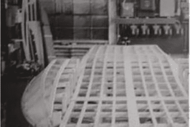 Black and white image of a large, grid-like structure under construction in a workshop, surrounded by various tools and materials.