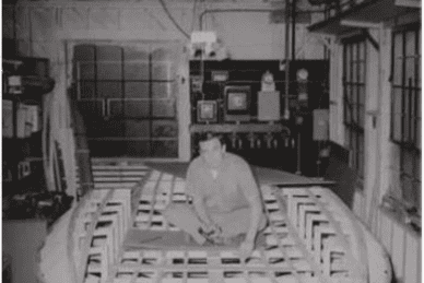 A person sitting on a large wooden structure frame in an industrial workshop with tools and machinery in the background.